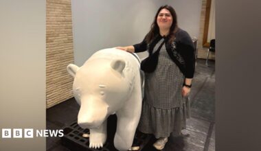 Becky Shepperson in a long dress and cardigan, smiling with her hand on a bear sculpture. The bear is white as it has not been painted yet.