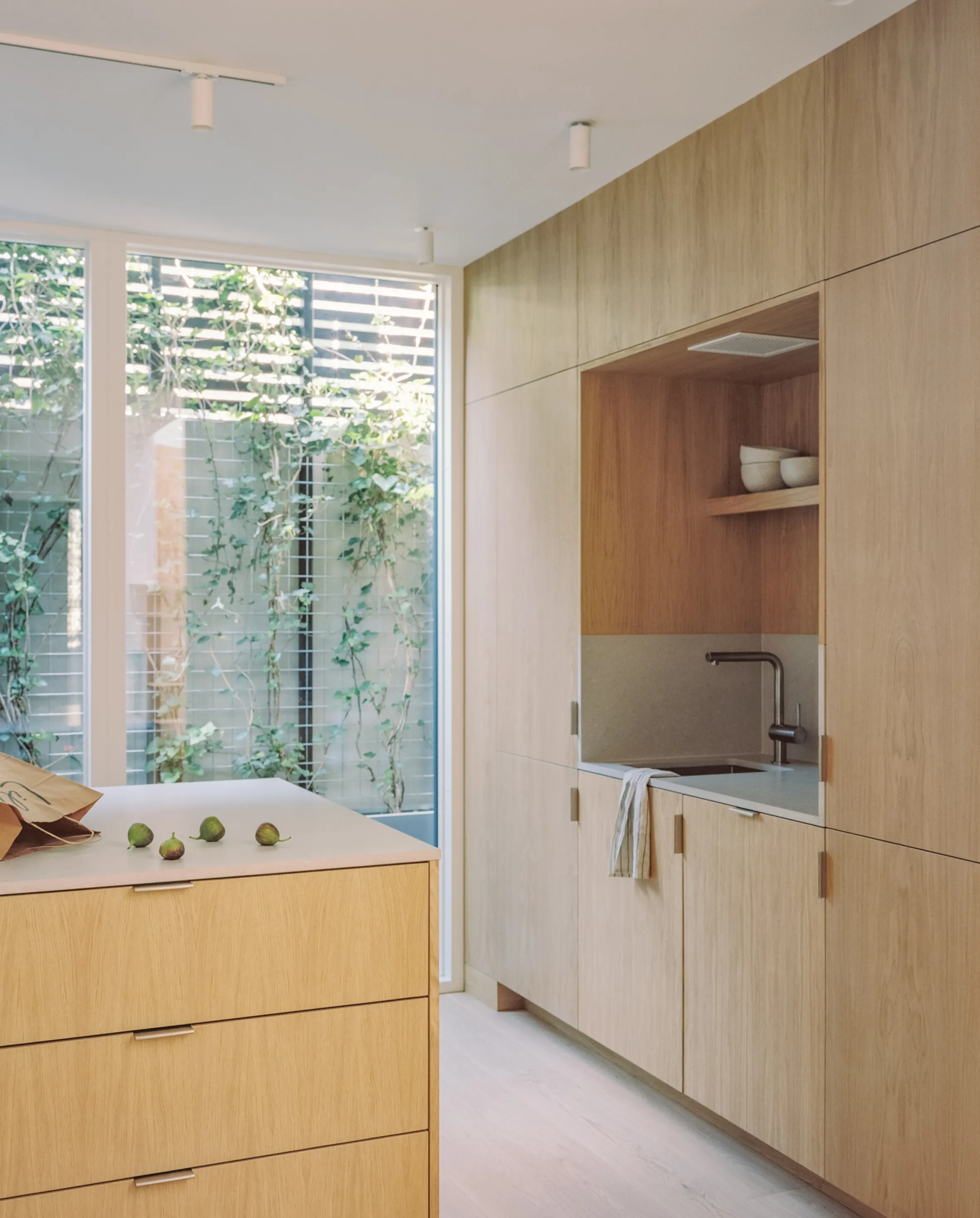 A kitchen with light wood cabinetry, a white countertop with a sink and faucet, and a large window looking out onto a trellis with green vines.