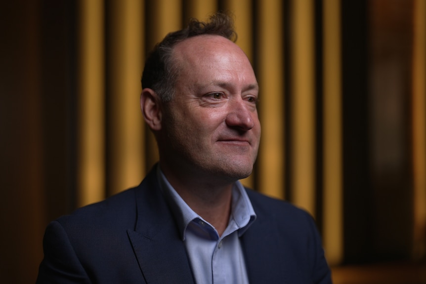 A middle-aged man wearing a navy suit jacket and light blue business shirt sits in a meeting room.