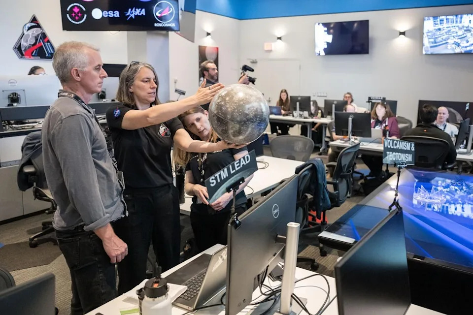 People in a control room discuss a space mission, holding a model planet. Screens and monitors surround them