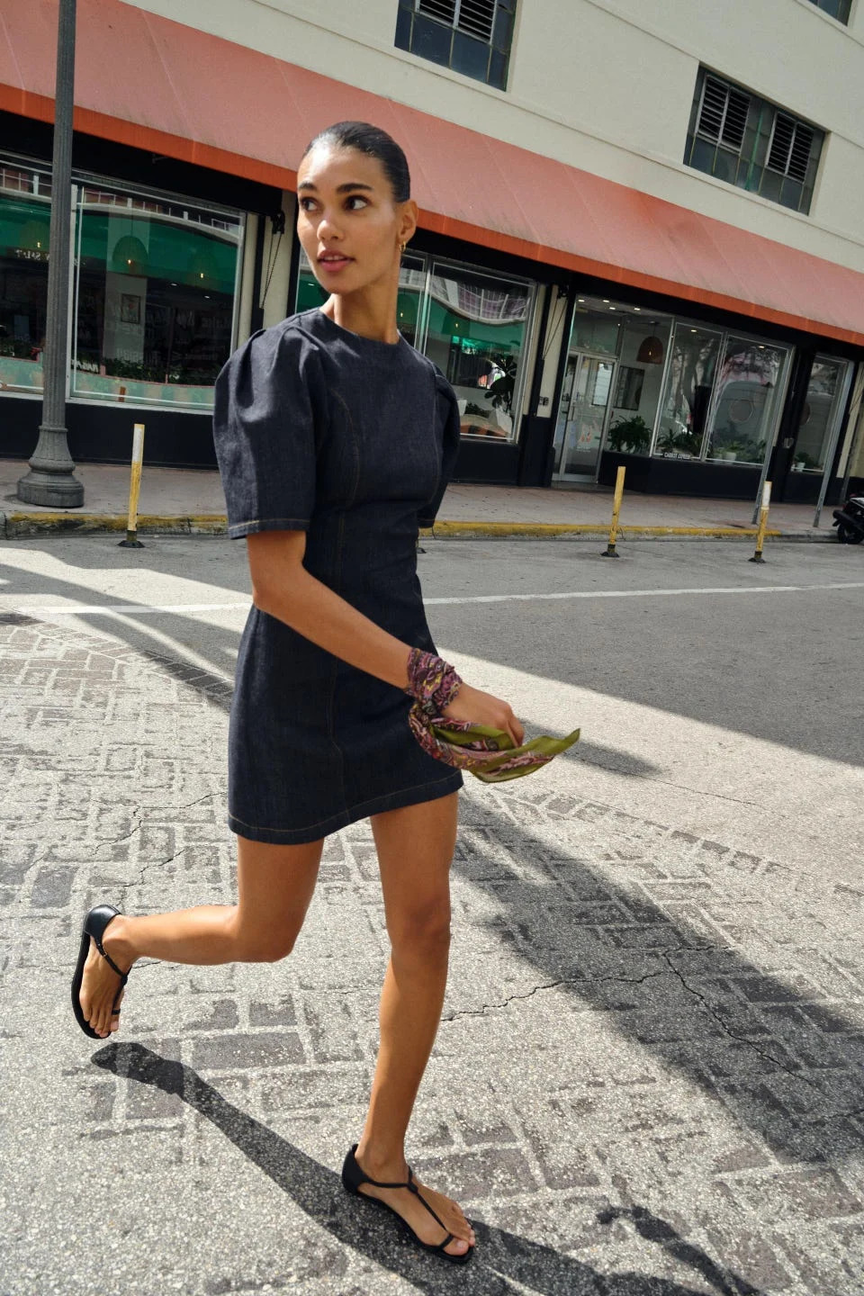 woman walking on a city street wearing a denim dress and sandals holding a scarf and a set of leaves