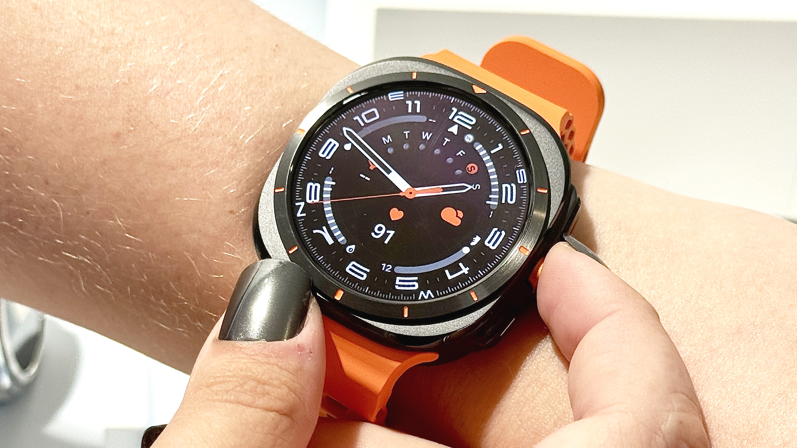 Close-up of the Samsung Galaxy Watch Ultra on a user's wrist with an analog watch face shown; the strap is bright orange