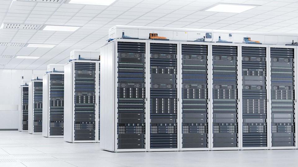 A wide shot of a pristine data center, showcasing several parallel rows of tall, white server racks filled with dark grey and black electronic hardware. The room has a white grid ceiling with rectangular lights and ventilation, and a light-colored tiled floor.