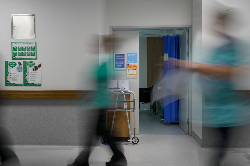 Blurred figures in hospital uniform walk through a hallway past an open room labelled BED 18