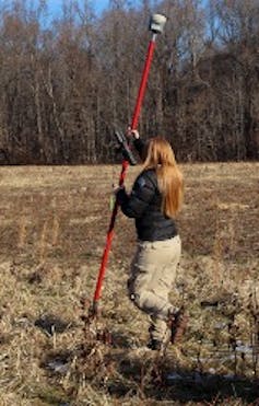 A woman holds a tall pole as she walks through a field with trees on one side.