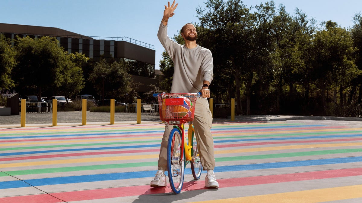 Steph Curry waving  on a rainbow bike in a street paved with rainbow lines.