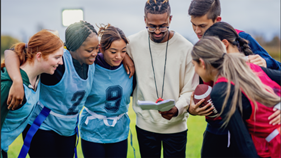 A coach holding a clipboard reviews plays with a diverse group of young athletes wearing flag football pinnies on an outdoor field
