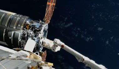 A large robotic arm reaches and grapples a silver-cylindric spacecraft with round solar arrays outside a space station.