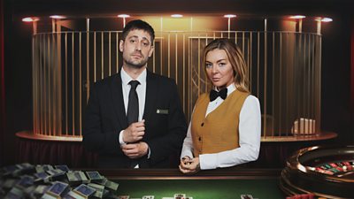 A man in a suit and a woman in a white shirt and yellow waistcoat stand by a casino table featuring a roulette wheel and a pile of cash. A cage is visible behind them.