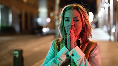 Sheridan Smith in character as Leanne. Stood on a city street at night. She clasps her hands together in front of her face, lost in thought.