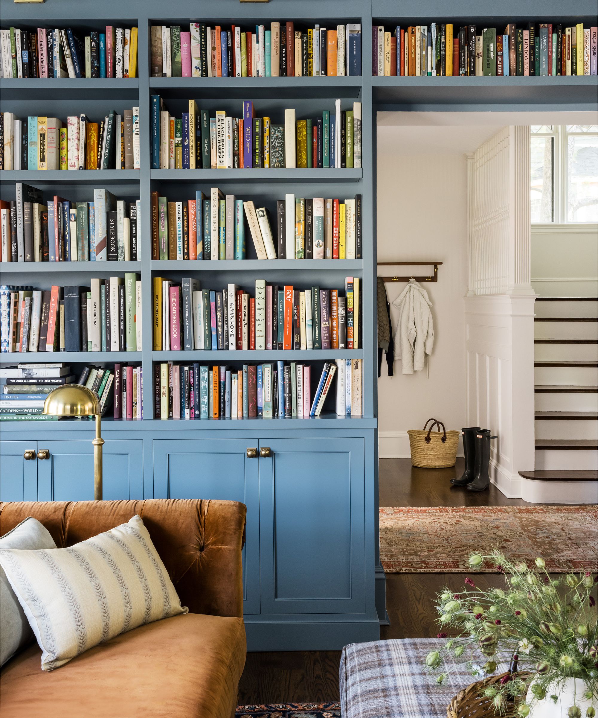 Living room with a blue-painted built-in bookcase, brown velvet sofa, plaid ottoman and gold floor lamp, looking through to a white entryway with brown wooden floor and coats hung on pegs