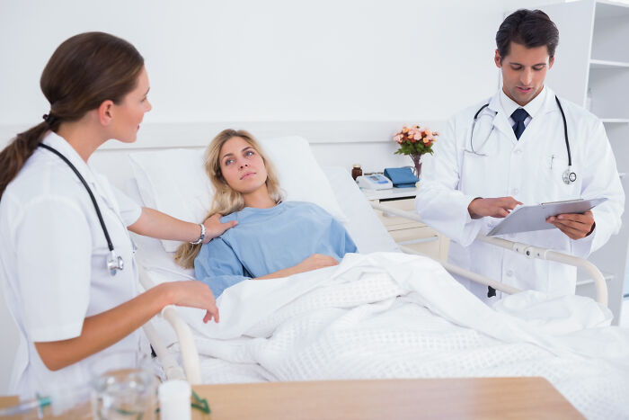 Two surgical staff attending a female patient in hospital bed during sedation, discussing patient confessions and care details.