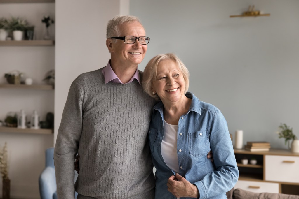 A serene senior couple, smiling and hugging in their living room, looking aside.