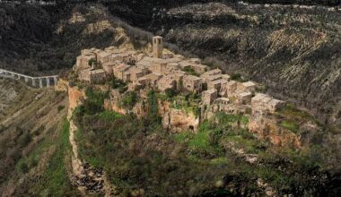 Aerial view of a small medieval village with stone buildings and a tall bell tower, perched atop a steep, rocky hill surrounded by green vegetation and deep valleys.