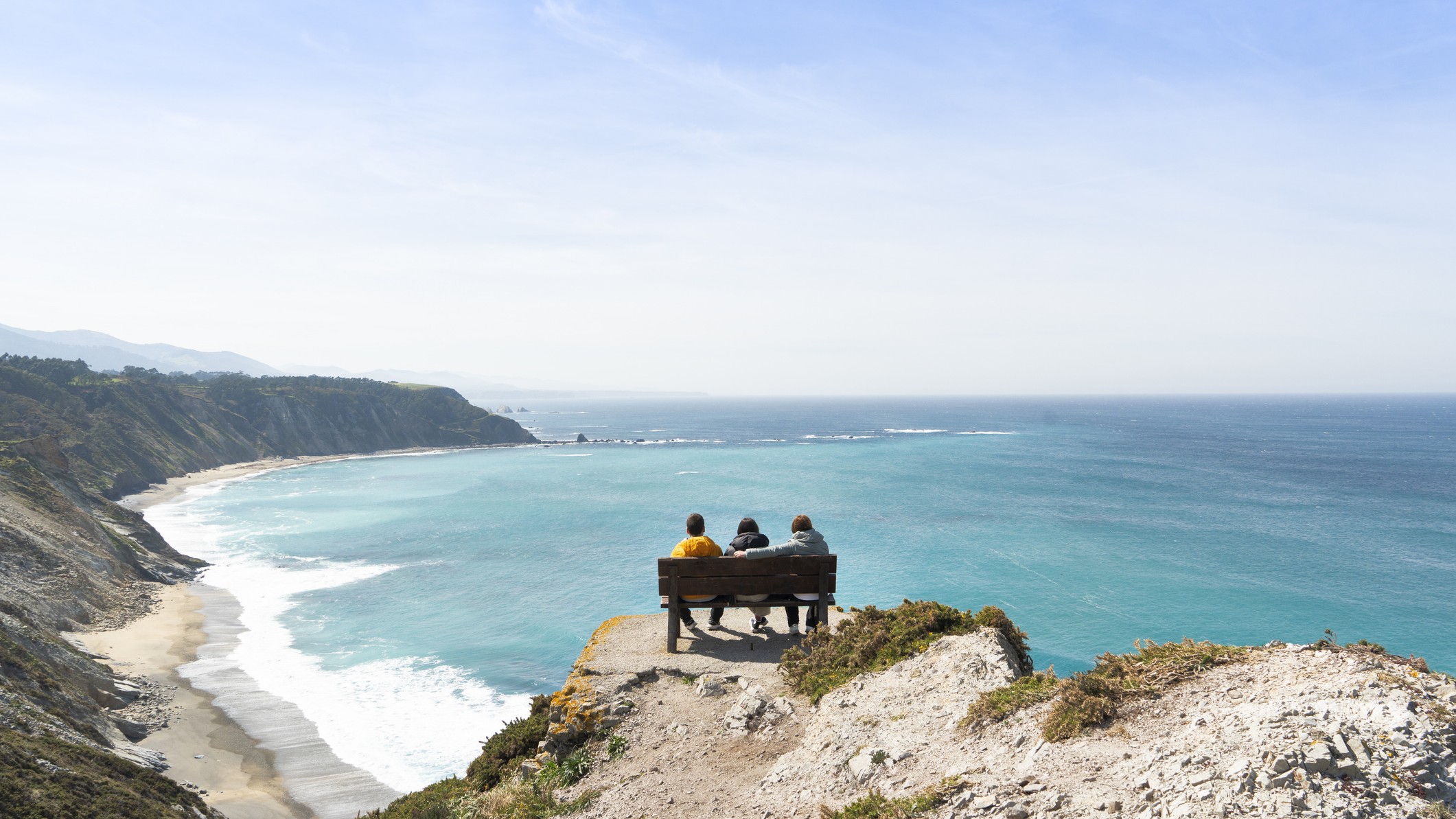 three people sit on a bench on the edge of a cliff with breathtaking views of the coastline.