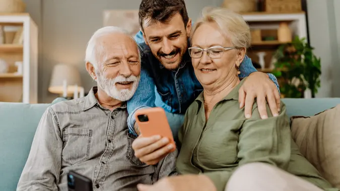 Younger Man leaning over his older parents sitting on the couch while they show pictures on a phone