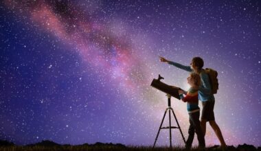 Two people with a telescope look up into a night sky that's full of stars and the Milky Way.