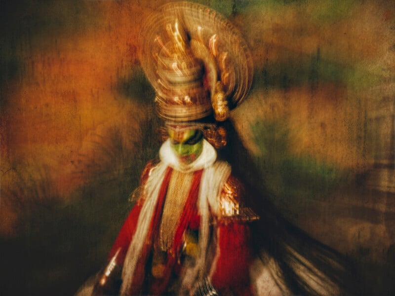 A blurred image of a Kathakali dancer in vibrant costume and elaborate headgear, with bold face paint and traditional attire, set against a warm, textured background.