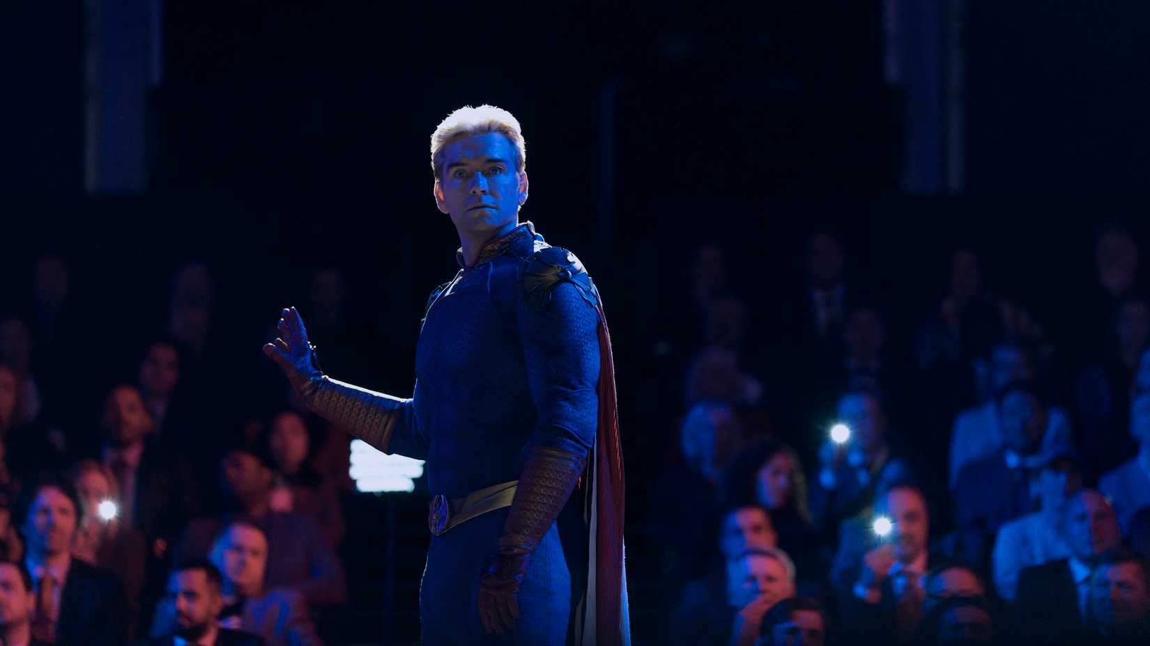 Homelander (Anthony Starr) stands with his back to an auditorium full of people, many holding cellphones to record video, in The Boys season 5