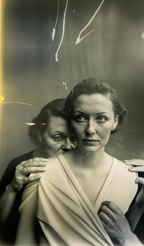 Two women pose together in a vintage-style, black-and-white photo. One woman stands behind, gently resting her hands on the other's shoulders and leaning in close, creating an intimate, mysterious atmosphere with light streaks and a hazy effect.