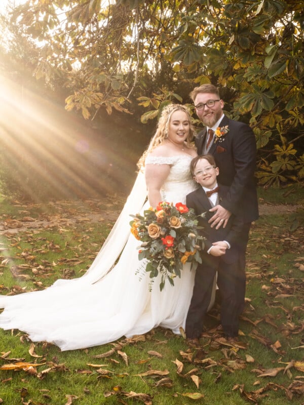 A bride in a white dress, a groom in a dark suit, and a young boy in a suit stand smiling together outdoors under a tree, with autumn leaves on the ground and sunlight streaming through the branches.