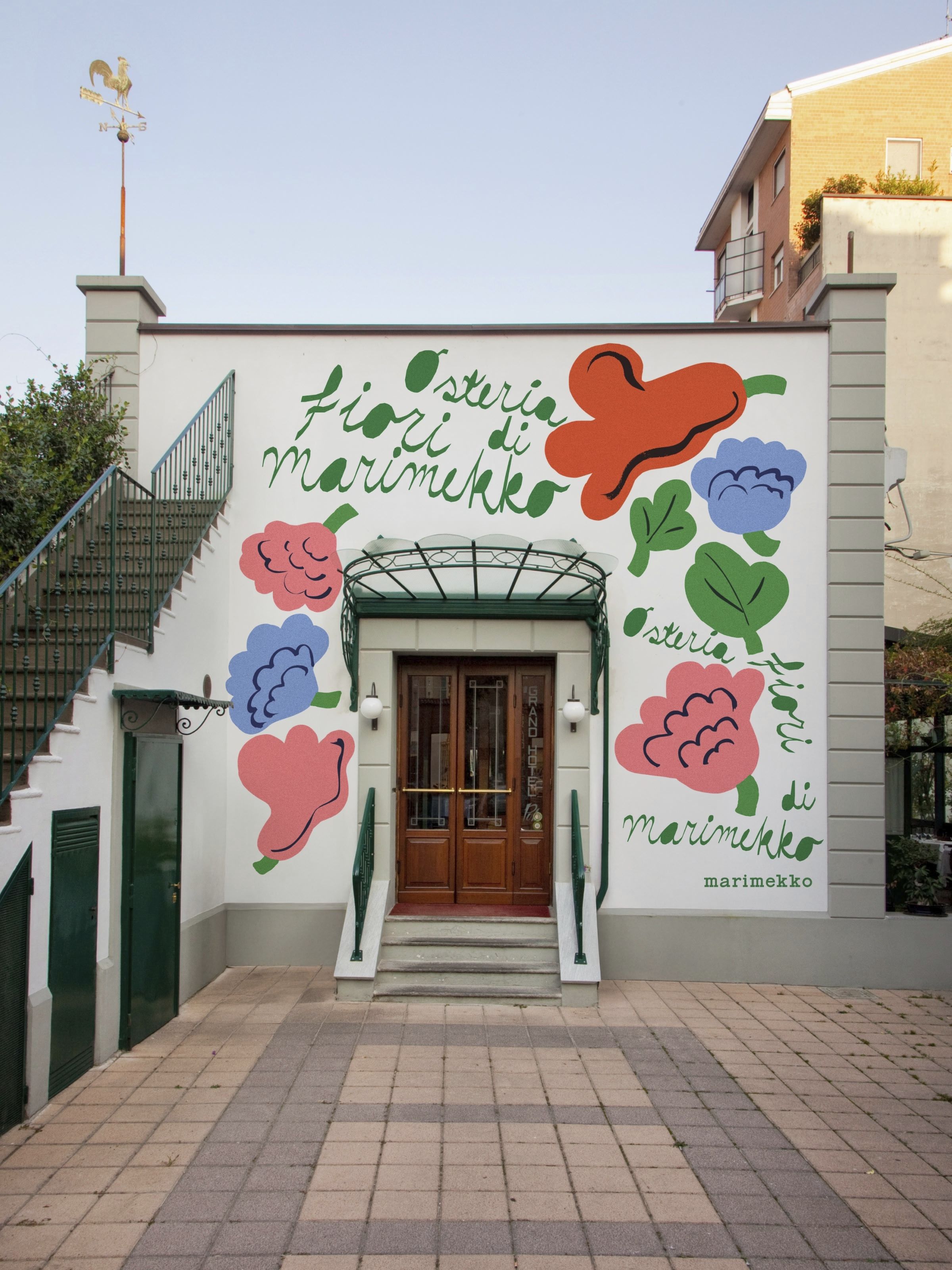 The exterior of an osteria, decorated with a floral mural, a table topped by floral-shaped serveware, and a woman dressed in a gray jumper and a white, brown, and blue skirt, handling yellow tuplis.