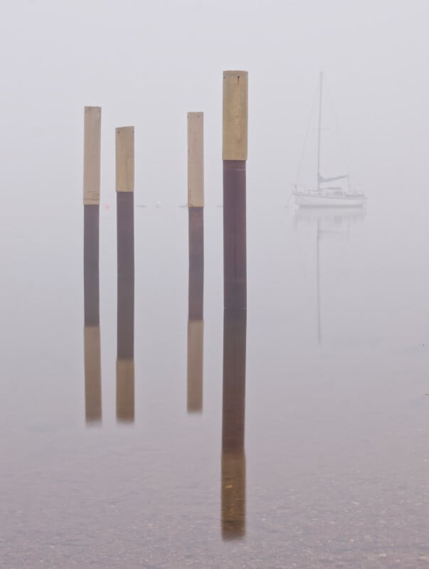 Four wooden poles stand in calm, shallow water with their reflections visible. A sailboat appears in the background, partially obscured by thick fog, creating a minimalist and tranquil scene.
