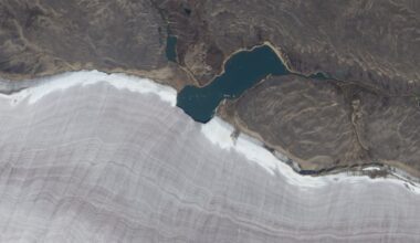 Satellite photo showing a lake bisecting the snowy edge of a gray ridged glacier