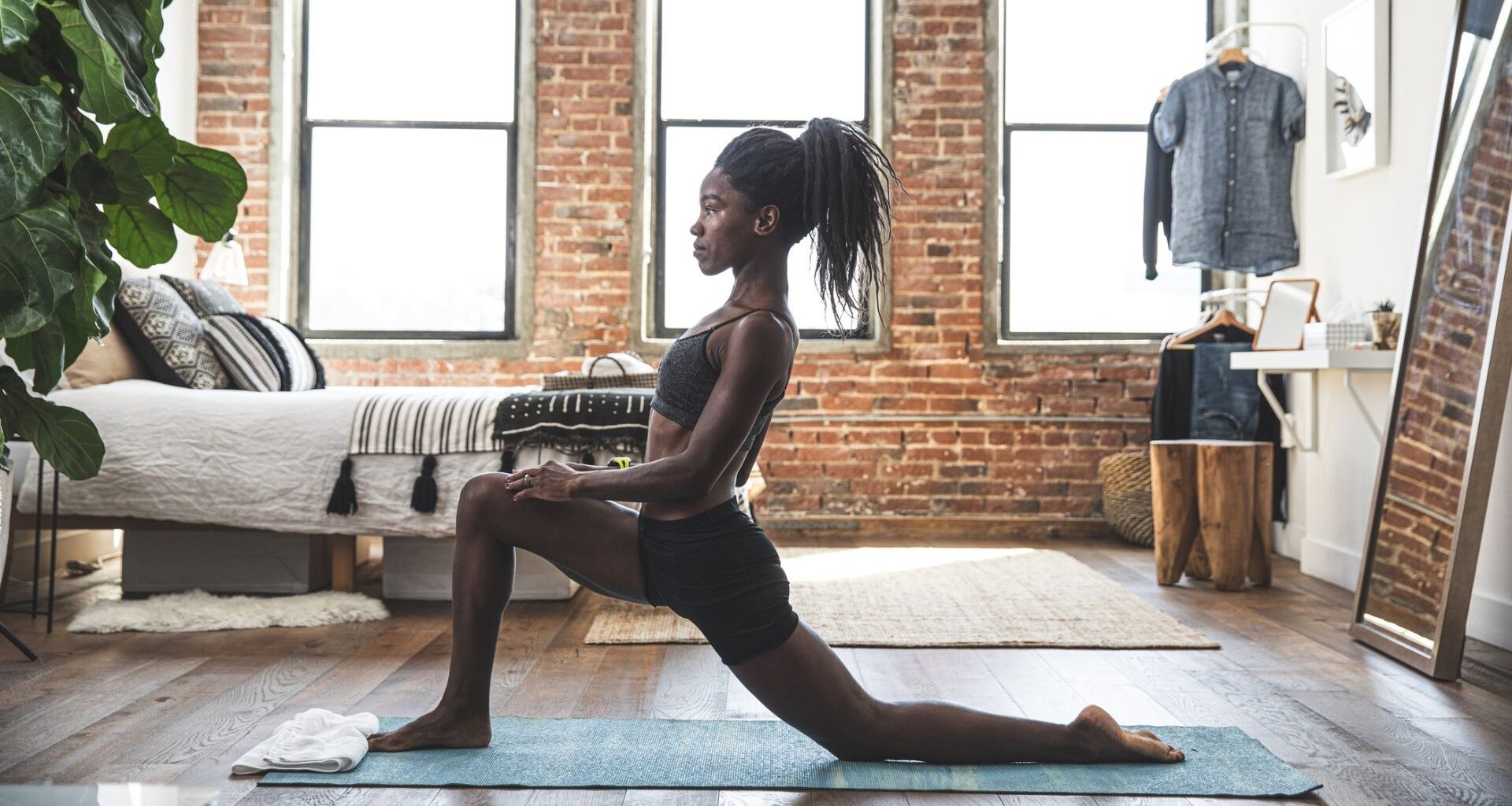 woman on an exercise mat performing a hip flexor stretch. she's sideways to the camera in a living room setting on wooden floors and exposed brick walls and windows behind her