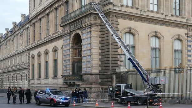 A ladder attached to a truck stands next to a large beige and grey building. A police car and several police officers are beside it.