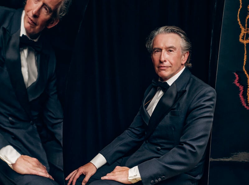 A man in a black tuxedo sits in front of a black backdrop, looking at the camera. His reflection is visible in a mirror beside him, showing his upper body and face from a different angle.