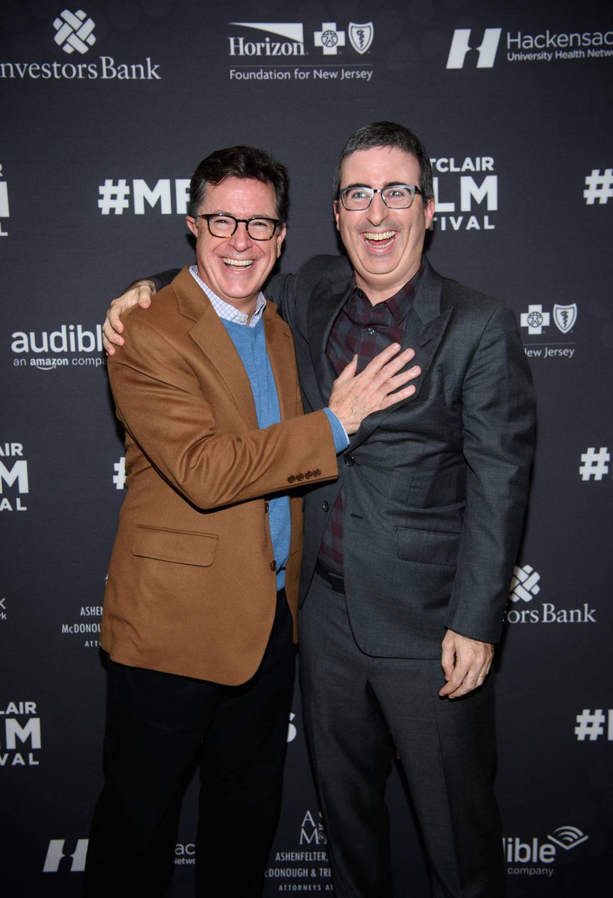 Two individuals, both wearing glasses and dressed in formal attire, are standing close to each other and smiling for a photograph against a backdrop adorned with event branding and hashtags.