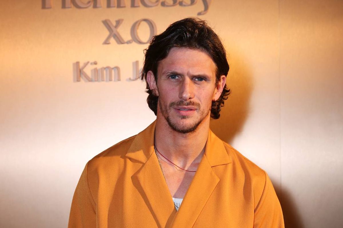 An individual is standing against a backdrop with a backdrop featuring text. The person is dressed in a yellow suit and appears to be posing for a photograph.