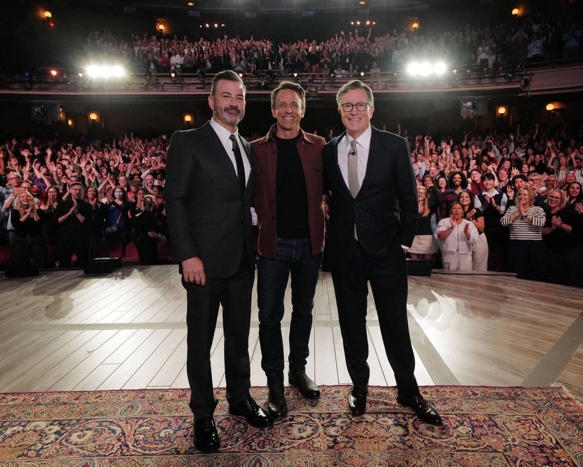 Three individuals in formal attire are standing on a stage in front of a large audience. The stage is adorned with a carpet, and the setting appears to be formal and possibly celebratory. The audience is seated in rows, and there is a sense of anticipation and engagement.