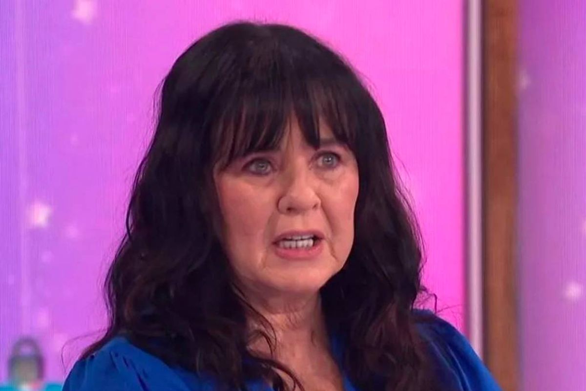 An individual with long, dark hair and a blue top stands against a vibrant pink background, appearing to engage in conversation or an interview.