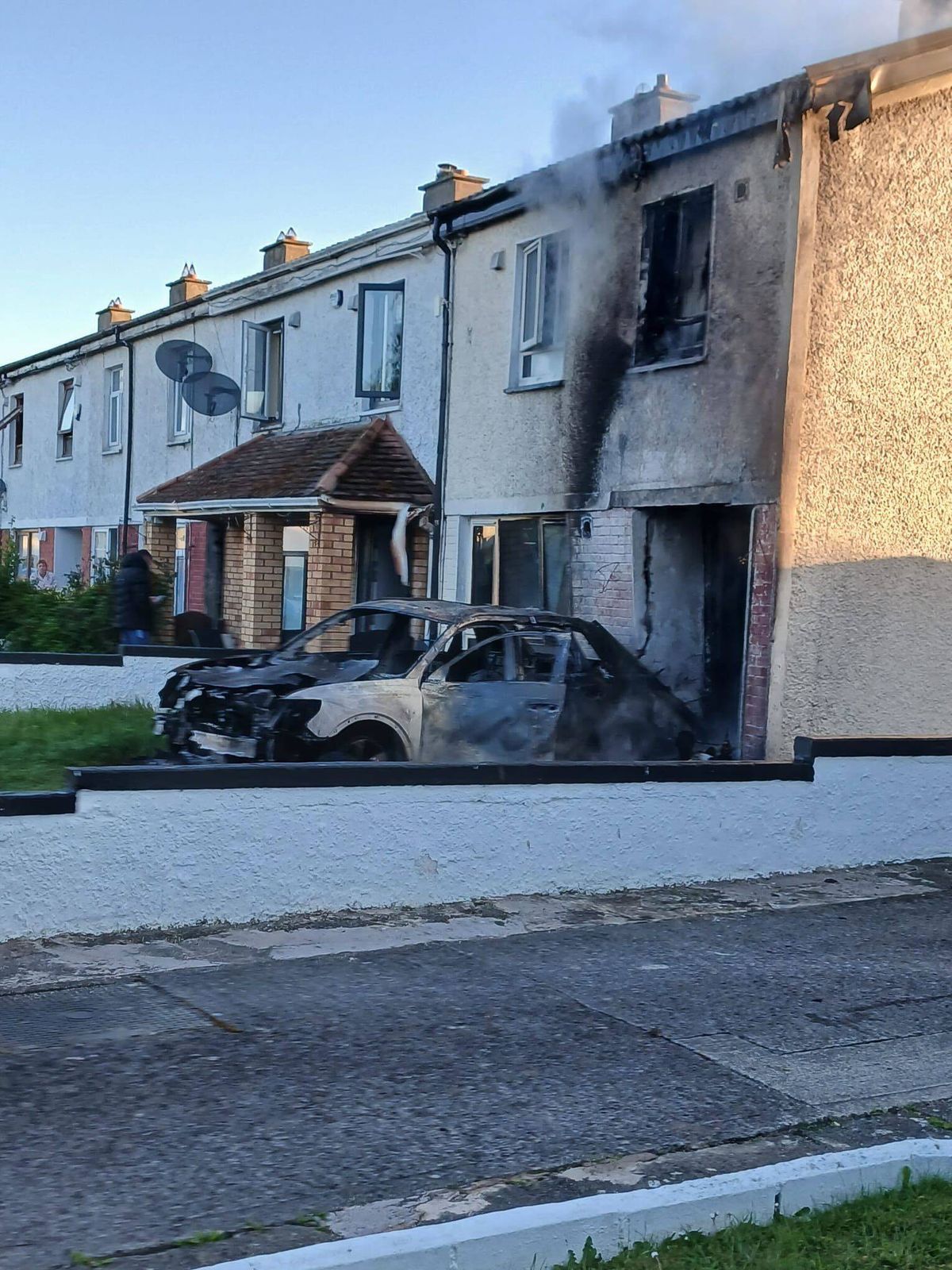 An image depicts a row of residential buildings with noticeable damage to one of the structures, characterized by soot and burnt marks. Several vehicles, including a black car, are parked in front of the damaged building, with one car appearing to have been severely affected by fire. The scene suggests recent damage or an incident involving fire.