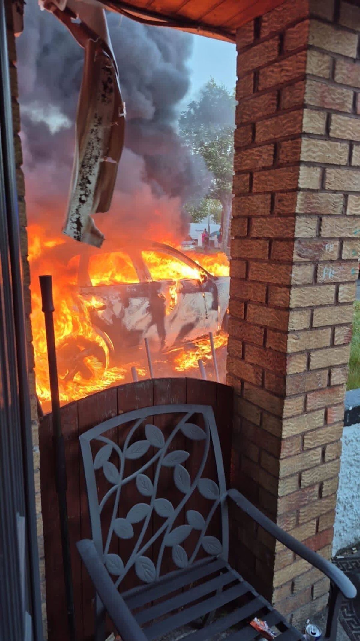 A vehicle engulfed in intense flames is visible through a glass door, with a brick column adjacent to the scene.
