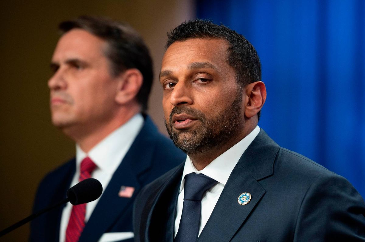 Two individuals in formal attire, one speaking into a microphone, with another individual standing behind him. Both individuals are wearing ties and are positioned in front of a blue backdrop.