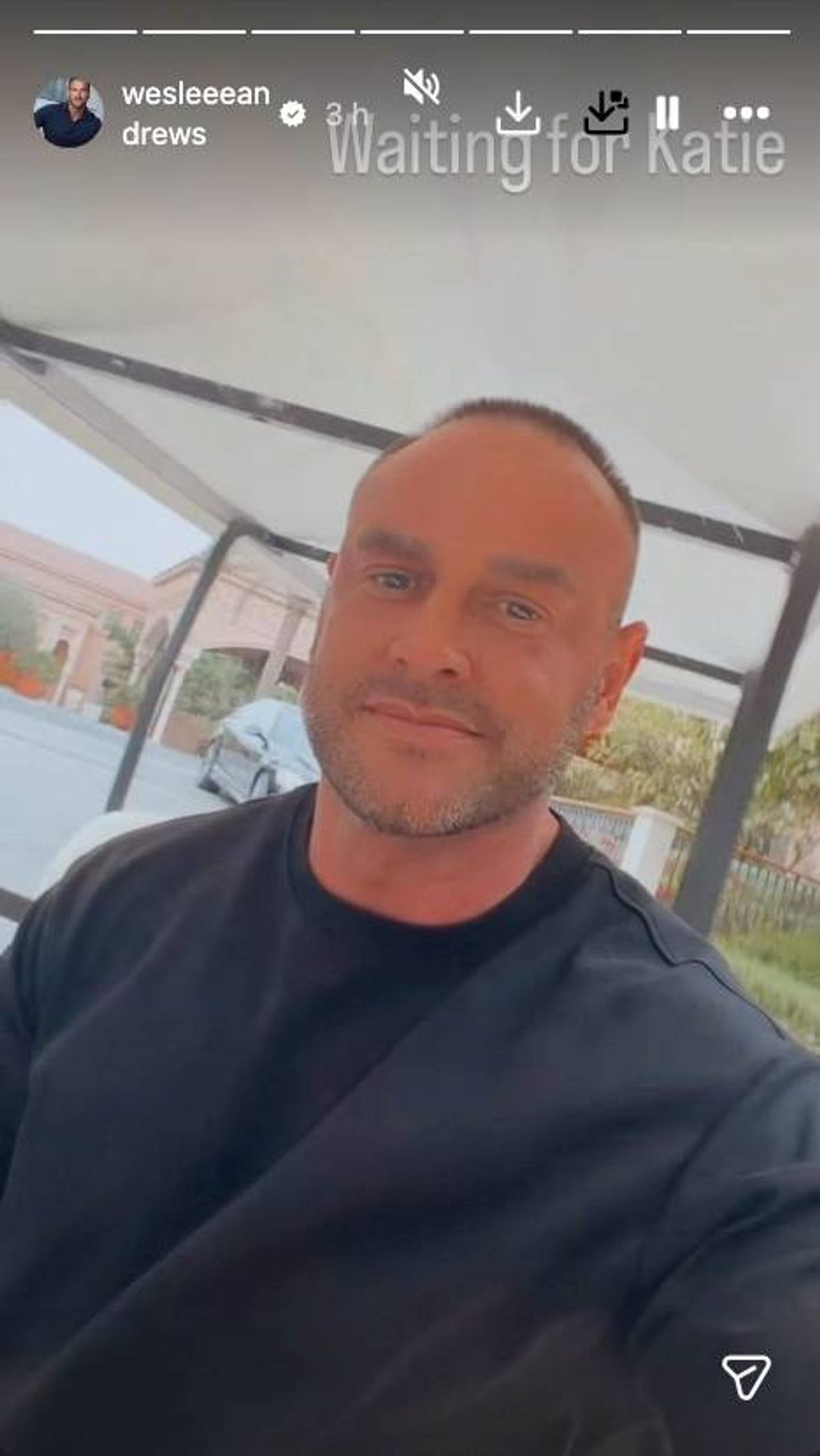 An individual with a bald head and a trimmed beard, dressed in a black t-shirt, is posing for a photograph with a contemplative expression against a backdrop of a building and a vehicle.