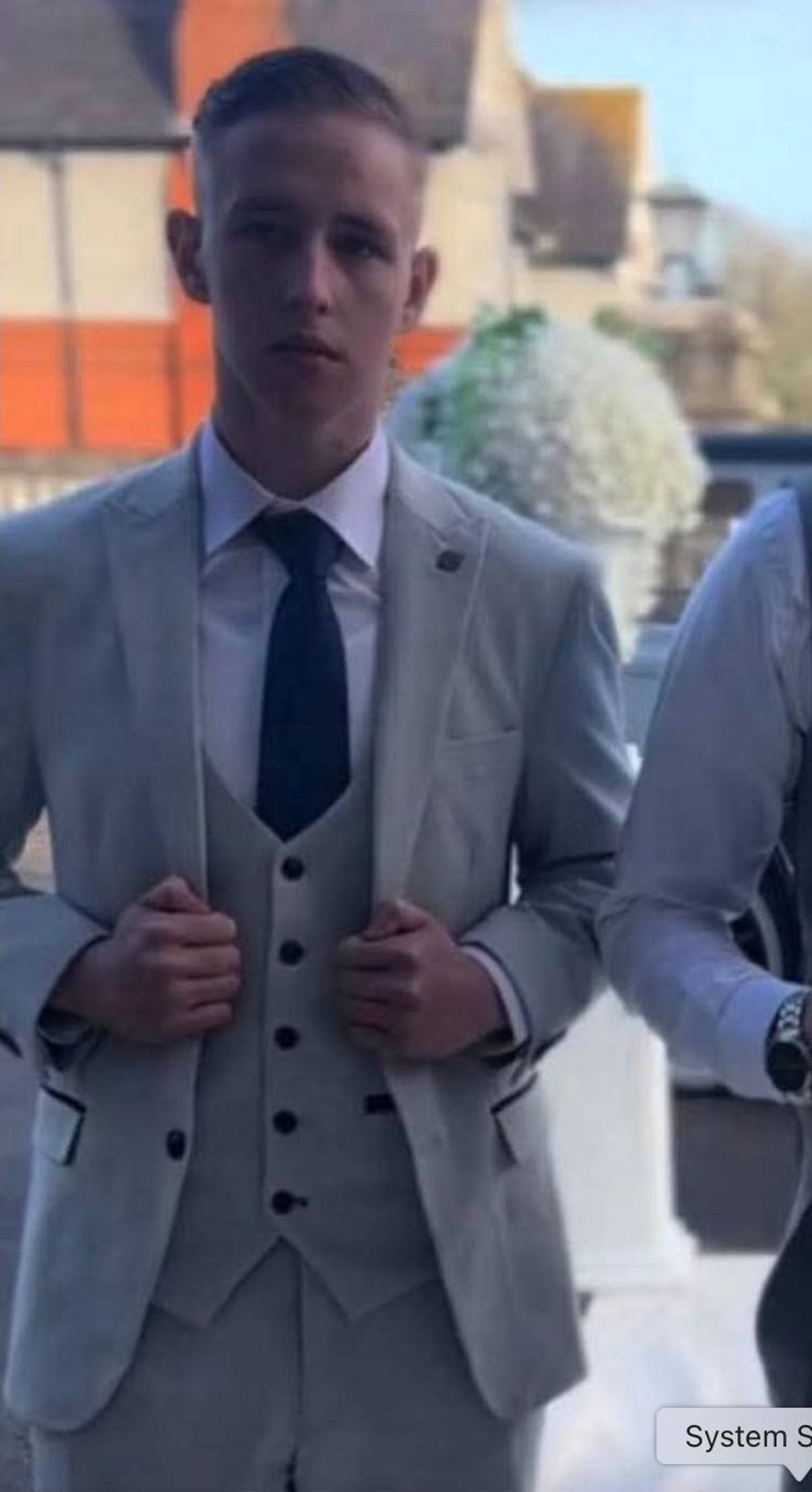 An individual dressed in a formal suit and tie is standing in front of an urban backdrop, possibly a building, with a focus on their contemplative expression.