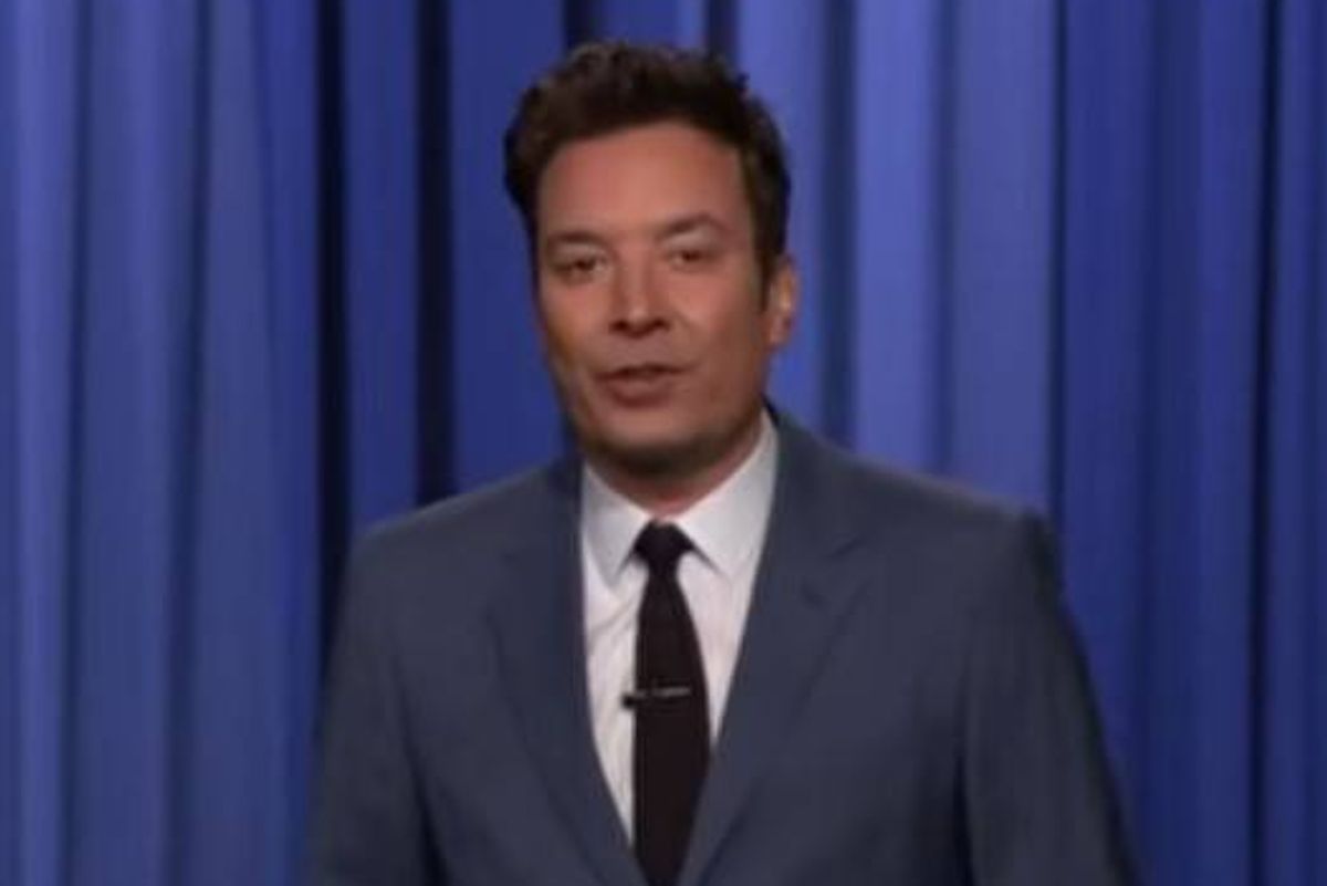 An individual dressed in a formal suit, consisting of a gray jacket, white shirt, and black tie, is standing in front of a blue curtain. He appears to be engaged in a formal presentation or speech, maintaining a serious expression.