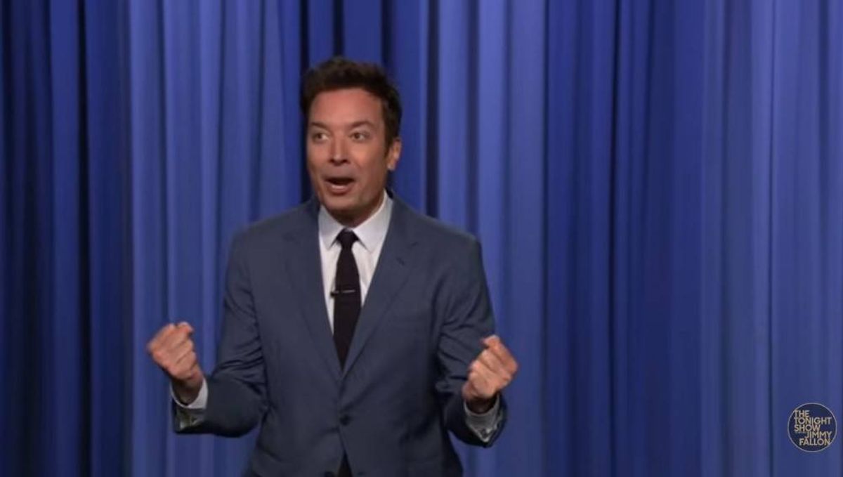 An individual dressed in a formal suit stands on stage, addressing an audience with a focused expression, positioned before a blue curtain.