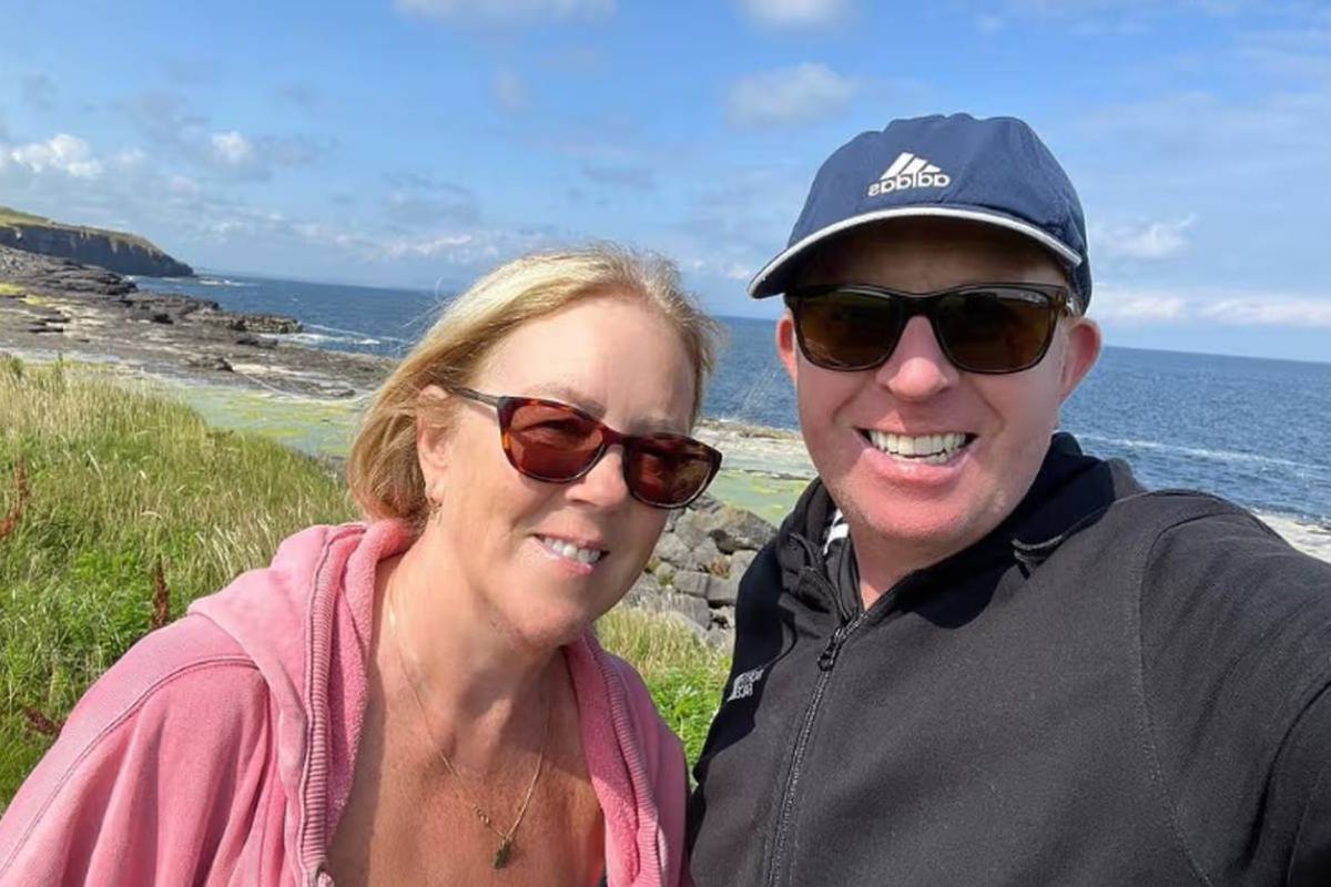 Two individuals are standing side by side in an outdoor setting, possibly on a coastal area. They are both wearing sunglasses and appear to be smiling. The person on the left is dressed in a pink jacket, while the individual on the right is wearing a black jacket. The background shows a grassy area with an ocean view and a partly cloudy sky.