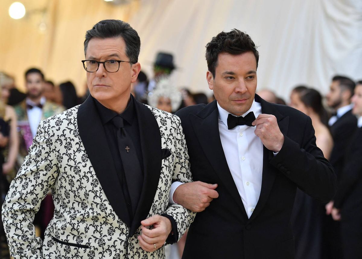 Two men in formal attire are walking together in a crowded setting, possibly attending a formal event. One man is wearing a patterned suit jacket with glasses, and the other is dressed in a classic tuxedo with a bow tie. In the background, a number of people in evening wear are visible, suggesting an upscale gathering.