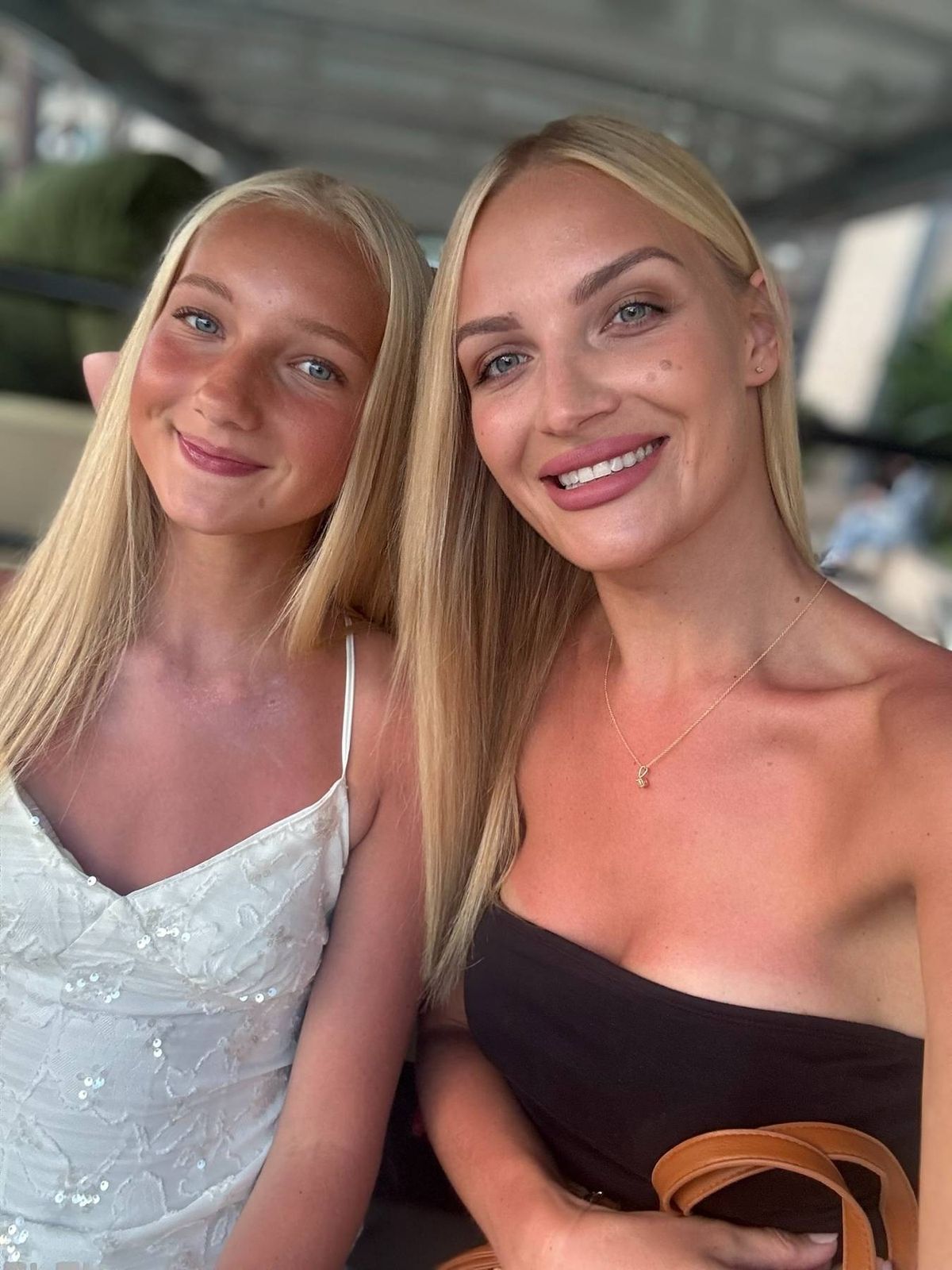 Two women with long blonde hair are standing next to each other and smiling for a picture. They appear to be in an outdoor setting with some architectural features in the background.