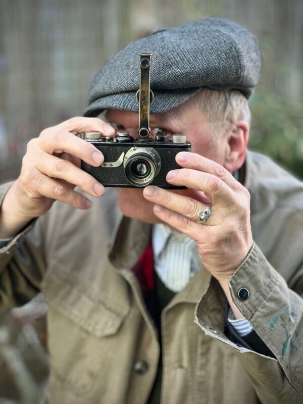An older person wearing a gray flat cap and beige jacket holds a vintage camera up to their face, focusing as if taking a photograph. The background is blurred.