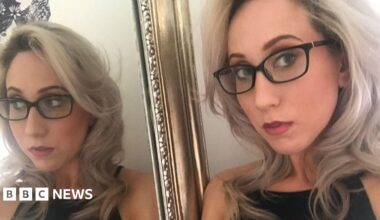 A woman with blonde hair and dark rimmed glasses stands next to a gold rimmed mirror which shows her full reflection