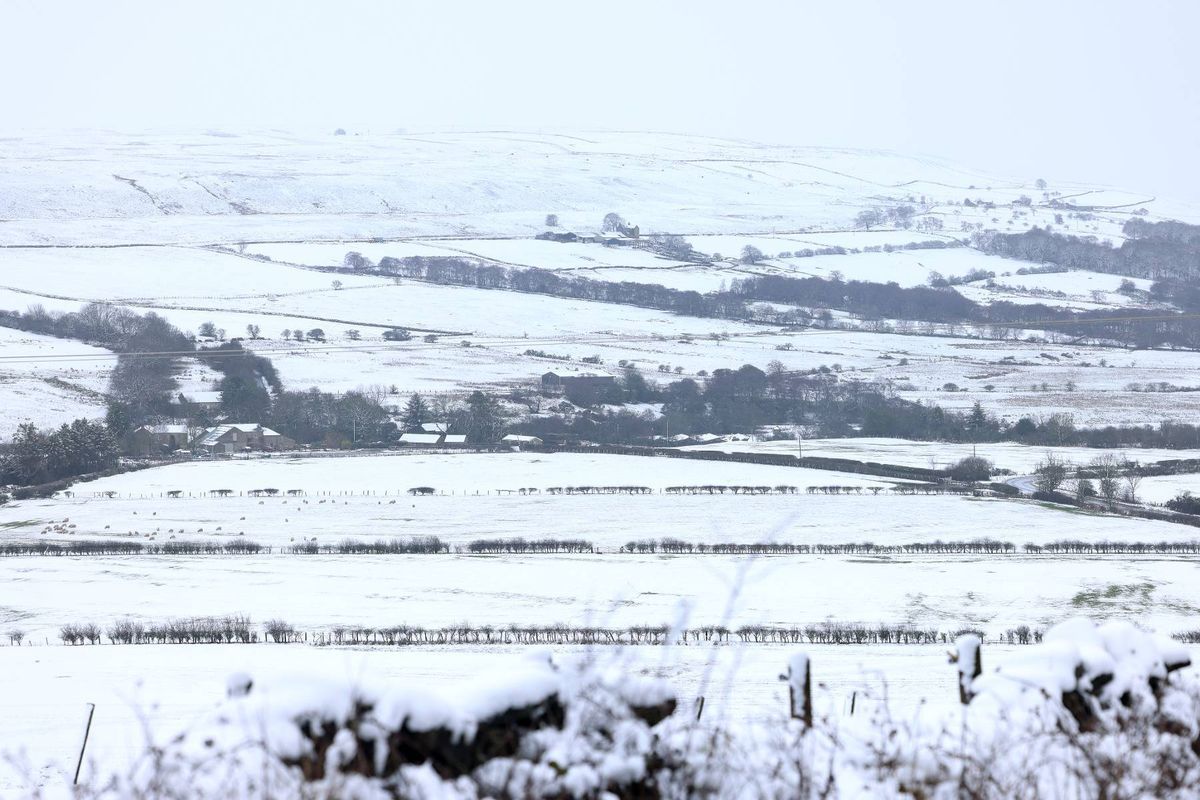 An expansive snowy landscape featuring rolling hills and distant structures blanketed in snow, with vegetation partially covered and a muted, wintry atmosphere.