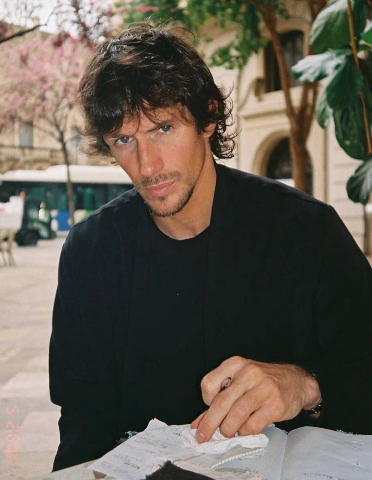 An adult male with short, curly hair, wearing a black t-shirt, standing outside in an urban setting. The background features a building with arched windows, trees, and a bus visible in the distance.