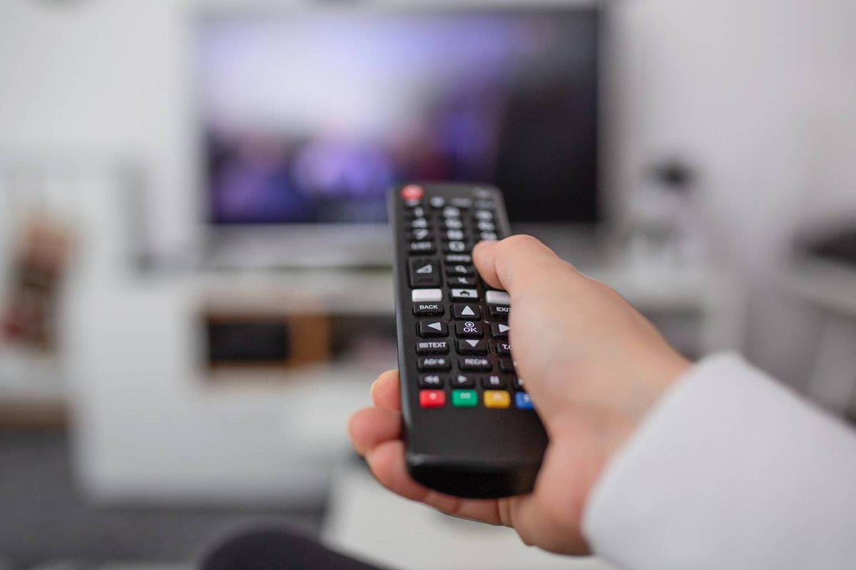 An individual is holding a modern television remote control, with a flat-screen television visible in the background, suggesting a scene of contemporary home entertainment.